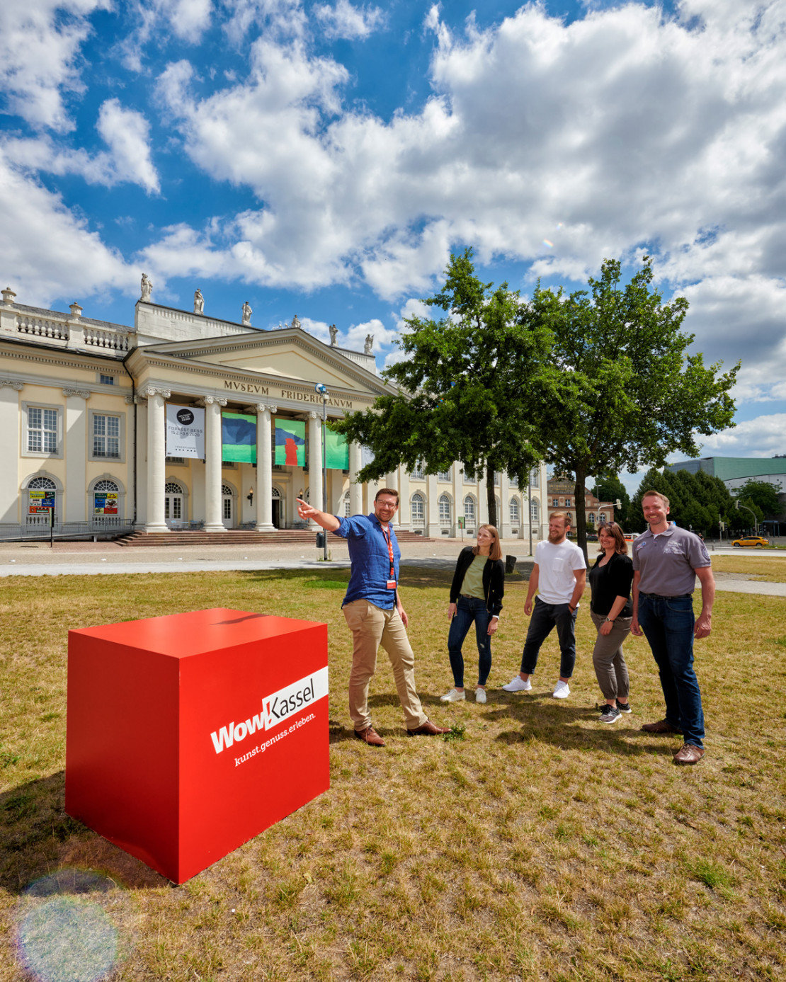 Eine Gruppe von fünf Personen stehen vor der Fridericianum Kassel mit einigen der Beuys-Eichen im Hintergrund und Wow-Kassel-Würfel im Vordergrund